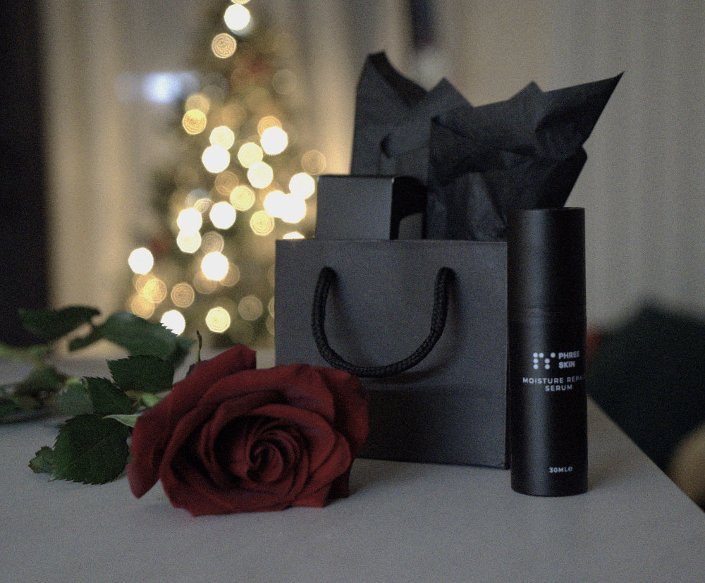 Black skincare product, red rose, and black gift bag on a blurred festive background